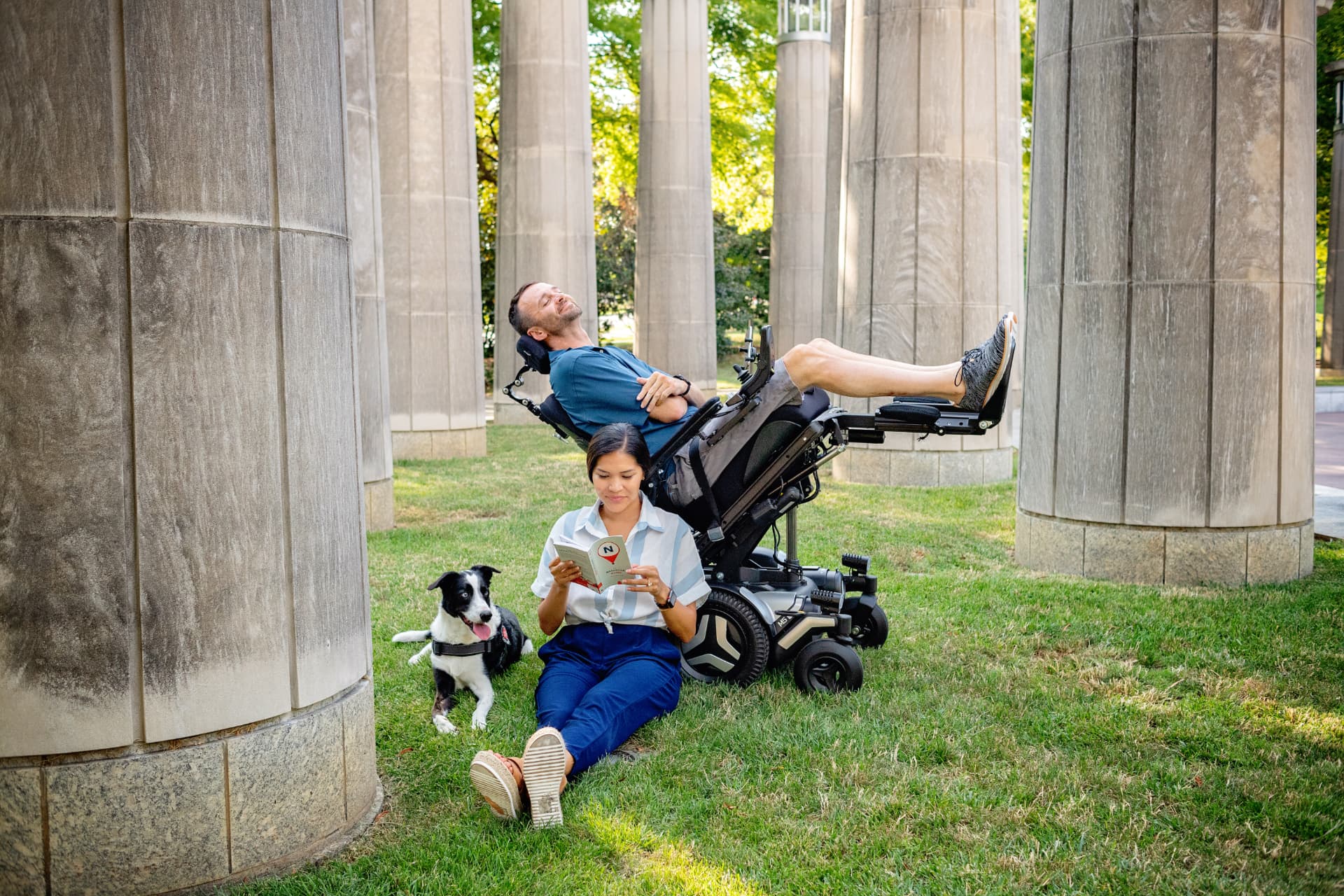 A man relaxing in a reclined power wheelchair in a park while a woman reads beside him with their dog